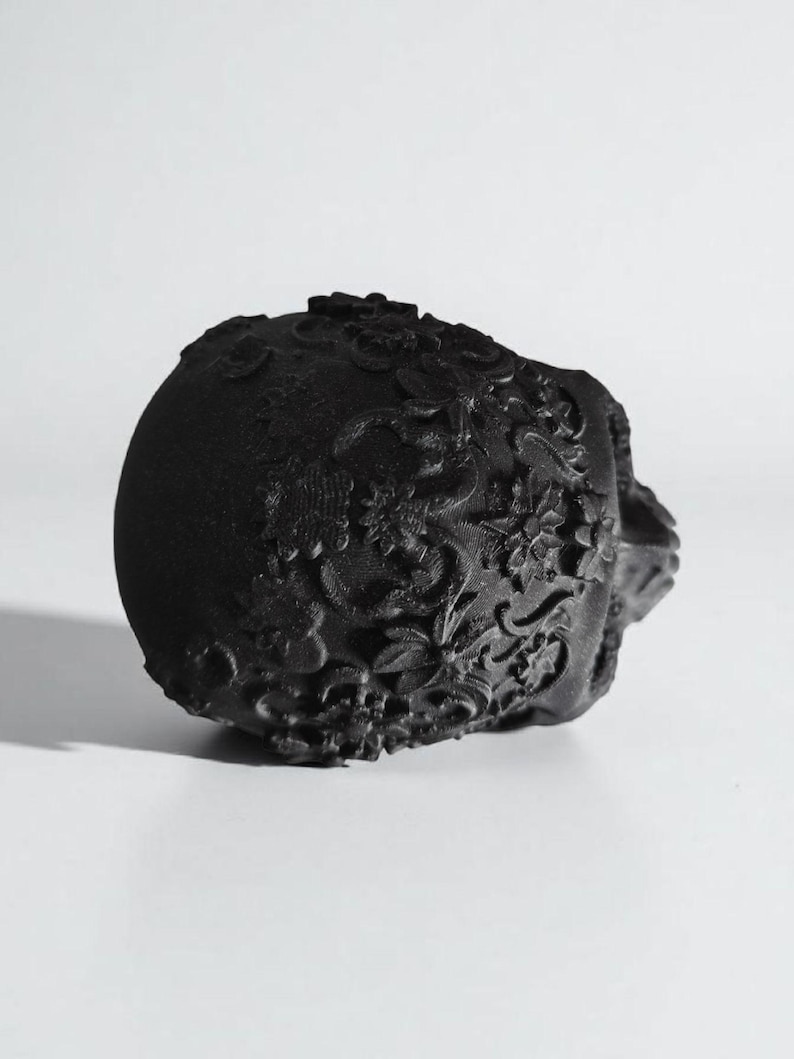 May include: Black decorative skull with floral patterns. The skull is made of a matte black material and features detailed, raised floral designs across its surface. The skull is positioned on a white background.