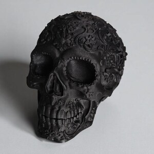 May include: A matte black decorative skull with intricate floral and scrollwork patterns. The skull features detailed facial features, including eye sockets, a nose cavity, and teeth. The design is a gothic or Day of the Dead theme.