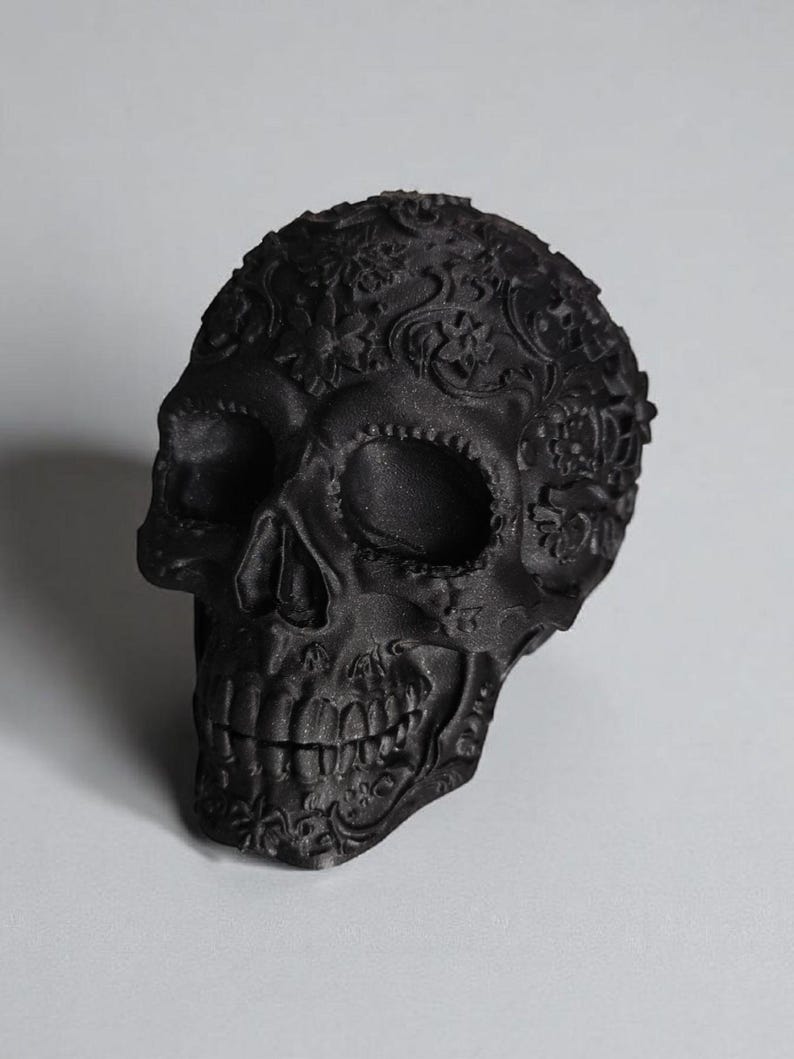 May include: A matte black decorative skull with intricate floral and scrollwork patterns. The skull features detailed facial features, including eye sockets, a nose cavity, and teeth. The design is a gothic or Day of the Dead theme.