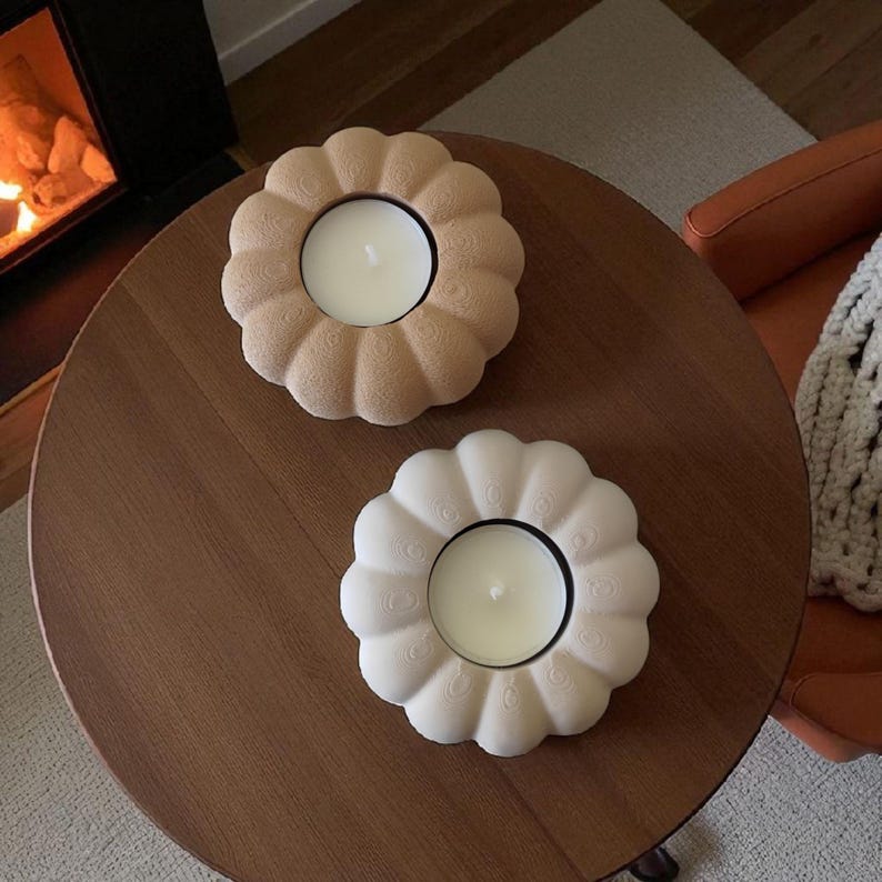 May include: Two flower-shaped candle holders with lit candles sit on a dark wooden table. One is beige, the other is white. The candle holders have a scalloped edge design. The background includes a fireplace and an orange chair.
