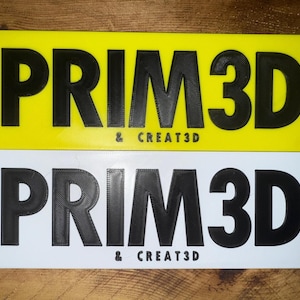 Pu&ograve; includere: Due cartelli rettangolari, uno giallo e uno bianco, ciascuno con il testo "PRIM3D & CREAT3D" in grassetto nero. I cartelli sembrano essere stampati in 3D.