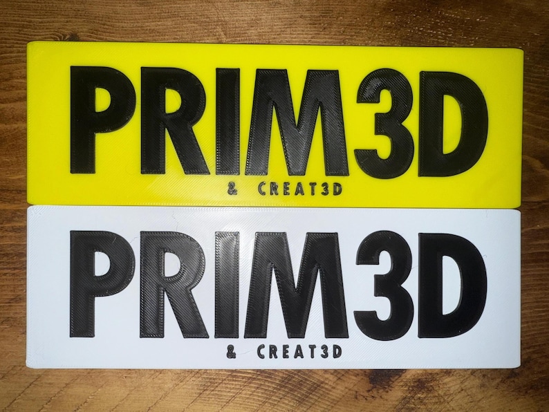 Pu&ograve; includere: Due cartelli rettangolari, uno giallo e uno bianco, ciascuno con il testo "PRIM3D & CREAT3D" in grassetto nero. I cartelli sembrano essere stampati in 3D.