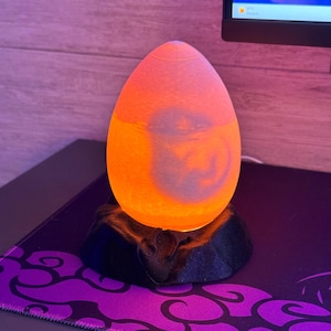 May include: An egg-shaped, orange and yellow lamp sits on a black base. The lamp is lit, and the light shines through the translucent material. The base is on a purple and black patterned surface. A computer monitor is in the background.