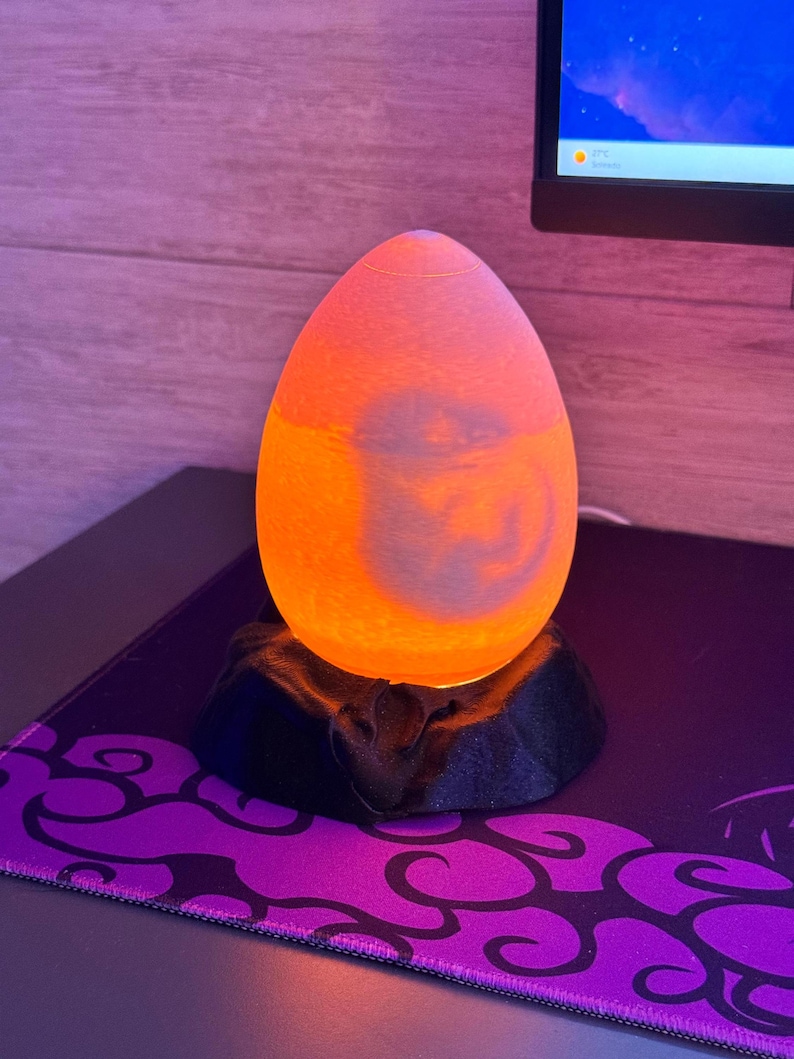 May include: An egg-shaped, orange and yellow lamp sits on a black base. The lamp is lit, and the light shines through the translucent material. The base is on a purple and black patterned surface. A computer monitor is in the background.