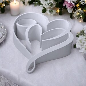 May include: A white heart-shaped decorative piece with a loop at the bottom. The design includes three cylindrical candle holders within the heart's outline. A lit candle and floral arrangements are in the background.