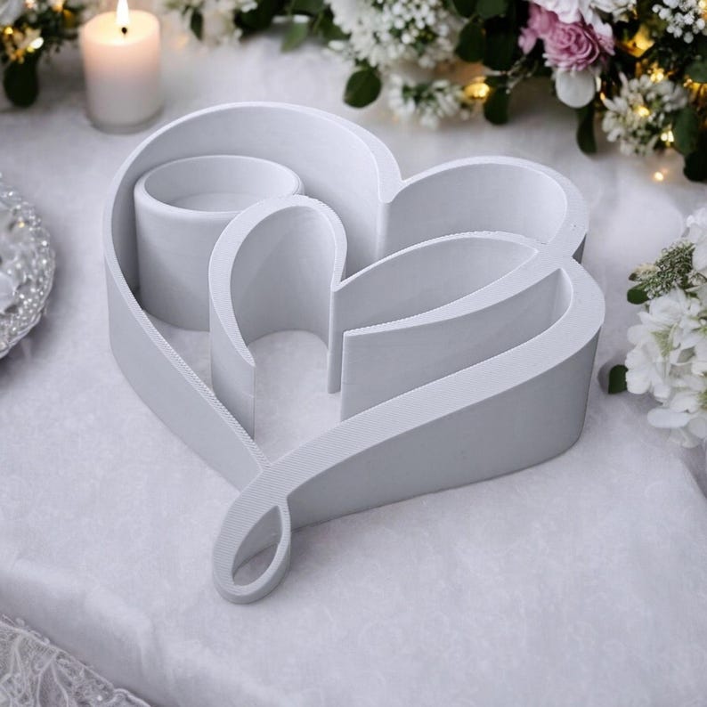 May include: A white heart-shaped decorative piece with a loop at the bottom. The design includes three cylindrical candle holders within the heart's outline. A lit candle and floral arrangements are in the background.
