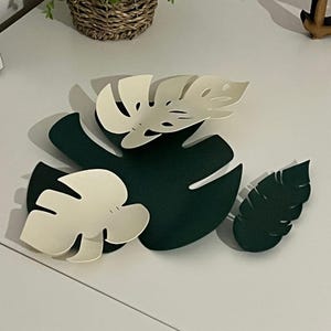 May include: A collection of paper cut-out Monstera leaves in shades of green and off-white. The leaves are arranged on a white surface, with a small woven basket and a plant in the background.