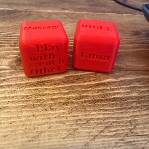 Puede incluir: Dos dados rojos con texto en relieve. Un dado dice "Massage" y "Play with each Other". El otro dado dice "1 min.". Los dados est&aacute;n sobre una superficie de madera.