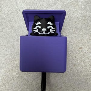 May include: A purple wall hook featuring a black cat design. The cat is smiling and peeking out of a purple box. The hook is black and extends from the bottom of the box. The design is 3D printed.