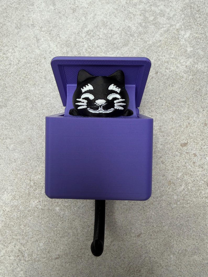 May include: A purple wall hook featuring a black cat design. The cat is smiling and peeking out of a purple box. The hook is black and extends from the bottom of the box. The design is 3D printed.