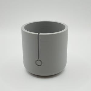 May include: A cylindrical, light gray object with a vertical slit and a circular indentation near the base. The object has a smooth, matte finish and a rounded bottom. The interior is hollow, creating a cup-like shape.