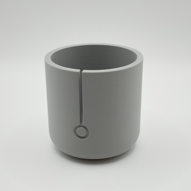 May include: A cylindrical, light gray object with a vertical slit and a circular indentation near the base. The object has a smooth, matte finish and a rounded bottom. The interior is hollow, creating a cup-like shape.