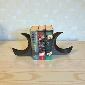May include: A dark gray, crescent-shaped bookend holds three books. The books have colorful covers with titles including "Harry Potter" and "Keeper of the Lost Cities." The bookend is made of a solid material and is designed to keep books upright.