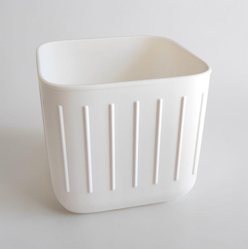 May include: A white, square-shaped storage container with vertical, indented lines on the sides. The container has rounded corners and a smooth, clean appearance. The interior is empty, suggesting it's ready for use.