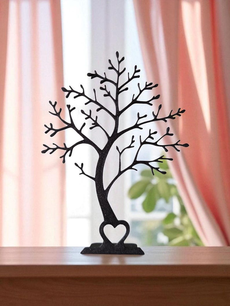 May include: A black metal jewelry tree shaped like a tree with a heart-shaped base. The tree has many branches for hanging necklaces, bracelets, and earrings. The background includes a window and pink curtains.