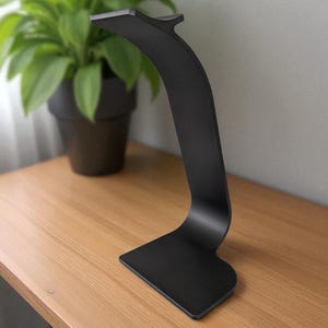 Peut inclure: Un support de casque noir &eacute;l&eacute;gant avec un design incurv&eacute;. Le support est en m&eacute;tal et poss&egrave;de une base plate et une section sup&eacute;rieure pour maintenir le casque. Il est pos&eacute; sur une surface en bois, avec une plante en arri&egrave;re-plan.