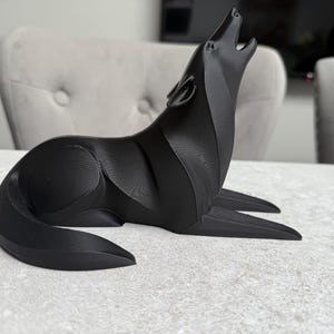 May include: A black, stylized wolf sculpture in a howling pose. The sculpture is made of a solid material and sits on a light-colored surface. The wolf's head is raised, and its mouth is open in a howl.