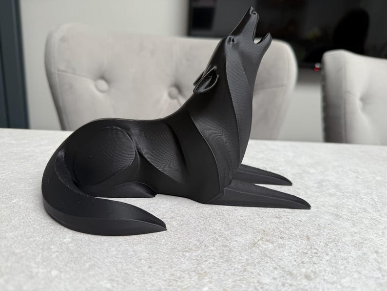 May include: A black, stylized wolf sculpture in a howling pose. The sculpture is made of a solid material and sits on a light-colored surface. The wolf's head is raised, and its mouth is open in a howl.