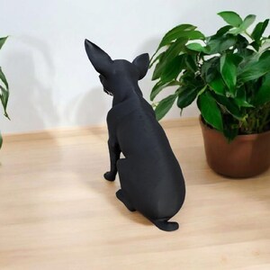 May include: A black dog figurine, viewed from the rear, sits on a light wood surface. The dog has large ears and is positioned near potted green plants. The figurine is a solid, matte black color.