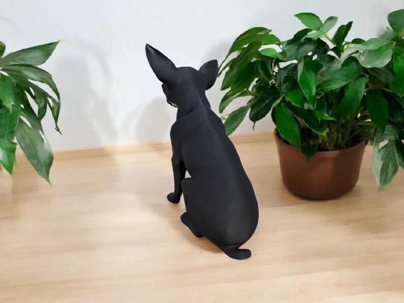 May include: A black dog figurine, viewed from the rear, sits on a light wood surface. The dog has large ears and is positioned near potted green plants. The figurine is a solid, matte black color.