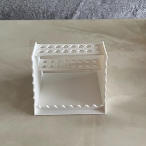 May include: A white, multi-tiered organizer with multiple circular holes and a scalloped edge. The organizer has two shelves with circular holes and a bottom shelf with a scalloped edge. The organizer is made of a solid white material.