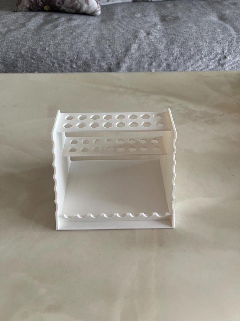 May include: A white, multi-tiered organizer with multiple circular holes and a scalloped edge. The organizer has two shelves with circular holes and a bottom shelf with a scalloped edge. The organizer is made of a solid white material.