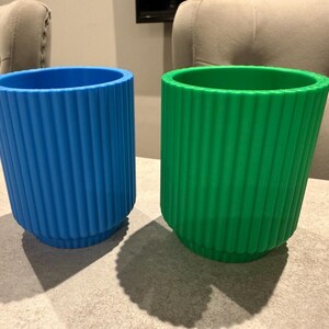 May include: Two ribbed, cylindrical containers. One is bright blue, the other is vibrant green. Both have a similar design with vertical ridges. The containers are sitting on a neutral-toned surface, with shadows cast below.