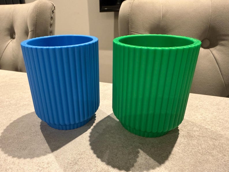 May include: Two ribbed, cylindrical containers. One is bright blue, the other is vibrant green. Both have a similar design with vertical ridges. The containers are sitting on a neutral-toned surface, with shadows cast below.