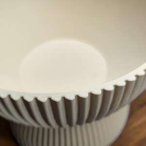 May include: Off-white, decorative bowl with a scalloped rim and vertical ribbed detailing. The bowl sits on a pedestal base, creating a tiered effect. The interior is smooth and the overall design is minimalist.