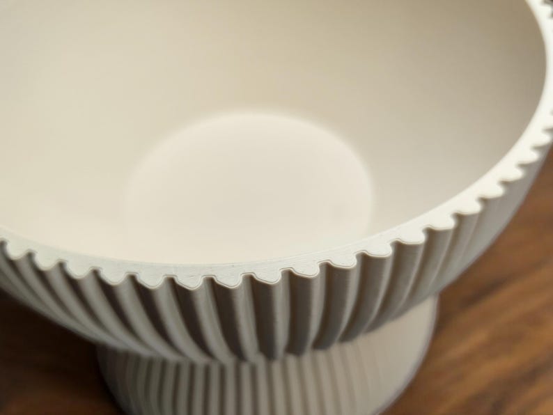 May include: Off-white, decorative bowl with a scalloped rim and vertical ribbed detailing. The bowl sits on a pedestal base, creating a tiered effect. The interior is smooth and the overall design is minimalist.