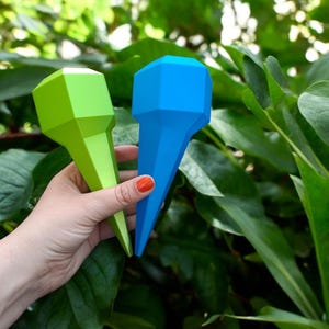 May include: Two geometric-shaped plant watering spikes, one green and one blue, are held against a backdrop of green foliage. The spikes have a faceted top and a tapered, pointed bottom, designed for insertion into soil.