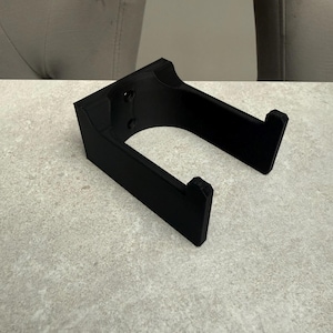 Op de afbeelding: Een zwarte, 3D-geprinte hoofdtelefoonstandaard. De standaard heeft een rechthoekige basis met twee schroefgaten en een gebogen opening. De armen zijn schuin en hebben kleine richels om de hoofdtelefoon vast te houden. Hij staat op een lichtgrijze ondergrond.