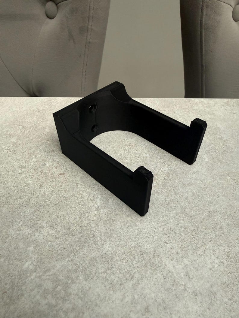 Op de afbeelding: Een zwarte, 3D-geprinte hoofdtelefoonstandaard. De standaard heeft een rechthoekige basis met twee schroefgaten en een gebogen opening. De armen zijn schuin en hebben kleine richels om de hoofdtelefoon vast te houden. Hij staat op een lichtgrijze ondergrond.