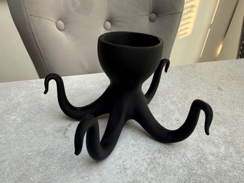 May include: A black octopus-shaped planter with a round opening and eight curved tentacles. The planter is made of a matte material and sits on a light grey surface. The tentacles are evenly spaced and curl upwards.