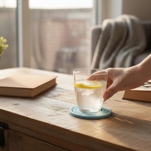 May include: A glass of water with a lemon slice on a blue coaster is held by a person. A book, eyeglasses, and a small potted plant sit on a wooden coffee table. A gray sofa and window are in the background.
