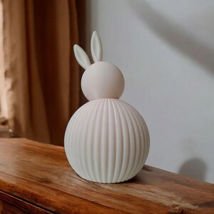 May include: A white ceramic bunny figurine with a ribbed body and two small ears. The figurine is sitting on a wooden surface.