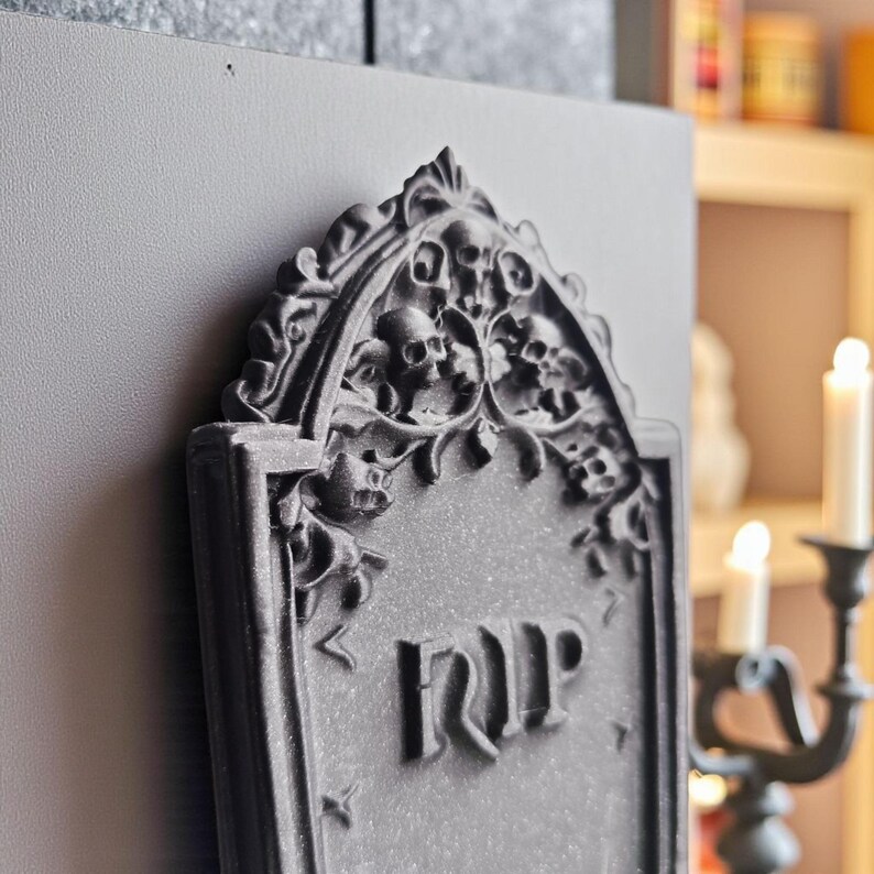 May include: A gray tombstone with the letters "RIP" carved into the front. The top of the tombstone features skulls and floral designs. The tombstone is set against a gray background, with a blurred background of a candle holder and books.