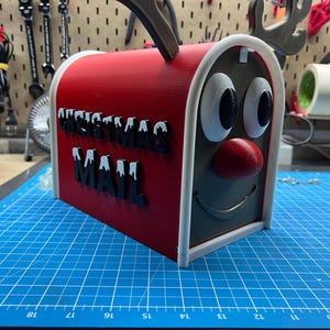 May include: A red and gray Christmas mailbox with a reindeer face design. The mailbox has the words "CHRISTMAS MAIL" in black letters with white accents. The mailbox has a red nose and antlers.
