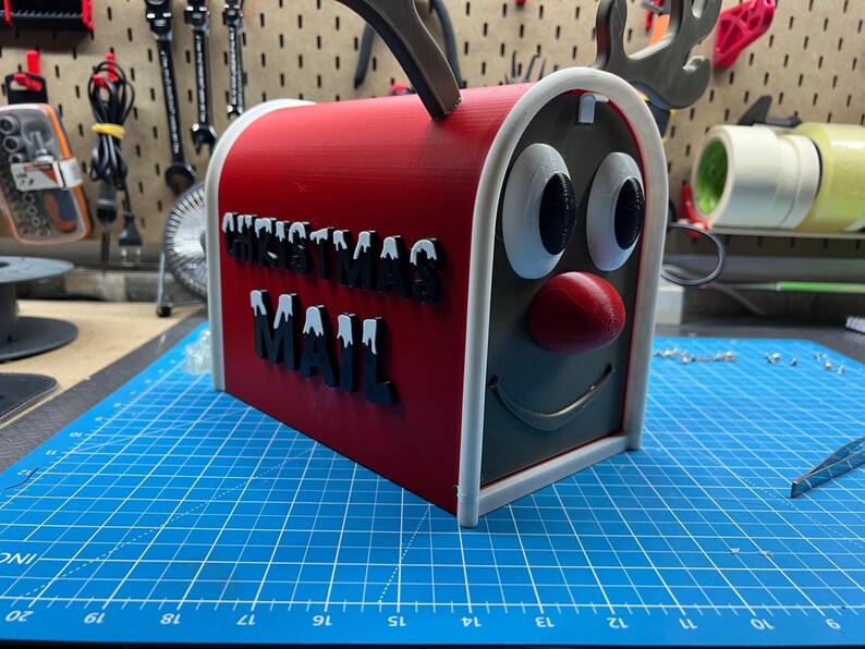 May include: A red and gray Christmas mailbox with a reindeer face design. The mailbox has the words "CHRISTMAS MAIL" in black letters with white accents. The mailbox has a red nose and antlers.