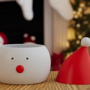 May include: A white snowman-shaped planter with black dot eyes and a red nose, next to a red Santa hat with a white pom-pom. The items are on a wooden surface, with a Christmas tree and Santa Claus in the background.