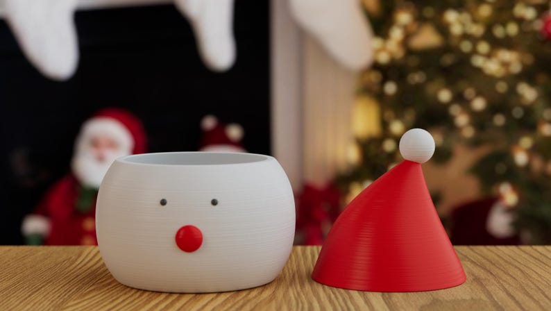 May include: A white snowman-shaped planter with black dot eyes and a red nose, next to a red Santa hat with a white pom-pom. The items are on a wooden surface, with a Christmas tree and Santa Claus in the background.