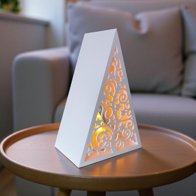 May include: White, triangular decorative lamp with a cut-out design on one side, revealing a warm, yellow glow. The lamp is placed on a round, wooden table. The lamp's design features intricate, swirling patterns.