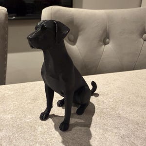 May include: A black dog figurine in a sitting position. The dog has a smooth, glossy finish and is positioned on a light-colored surface. The figurine is a decorative item, likely for home decor.
