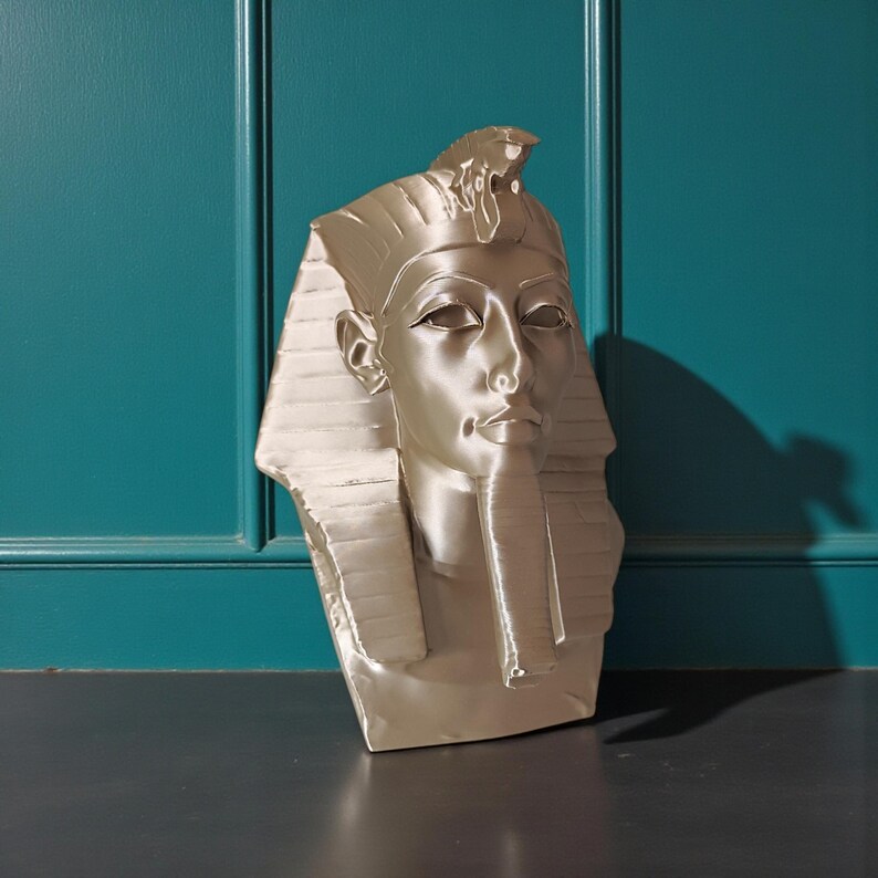 May include: A metallic gold bust of an Egyptian pharaoh, featuring a detailed headdress and serene facial expression. The sculpture is set against a teal wall and a dark surface, highlighting the intricate design and historical theme.