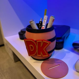 May include: A 3D-printed pen holder shaped like a barrel, featuring the letters "DK" in red with a gold outline. The barrel is peach-colored with black bands. Several pens and markers are inside. A matching coaster is next to the barrel.
