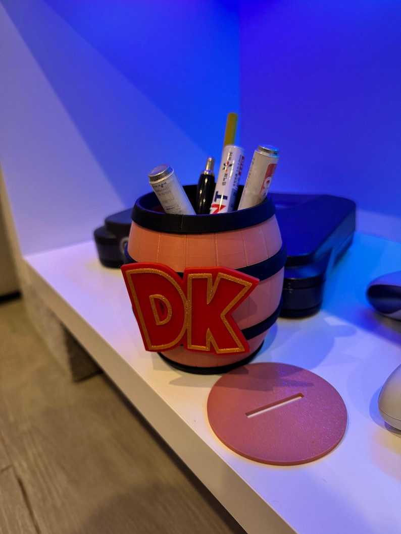 May include: A 3D-printed pen holder shaped like a barrel, featuring the letters "DK" in red with a gold outline. The barrel is peach-colored with black bands. Several pens and markers are inside. A matching coaster is next to the barrel.