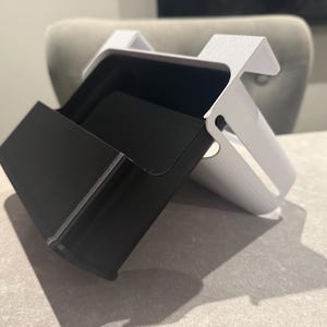 May include: A two-toned desk organizer with a black compartment and a white, angled support structure. The organizer is designed to hold items and has a modern, minimalist aesthetic. The black compartment is open and accessible.