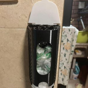 May include: A black plastic bag dispenser with a white lid is mounted on a beige tiled wall. The dispenser has a rectangular shape with a perforated design and a clear window showing the contents. The dispenser is filled with plastic bags.