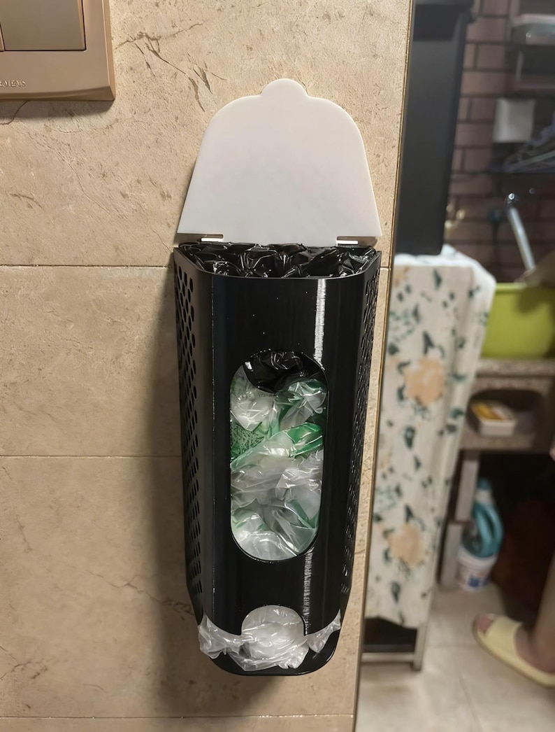 May include: A black plastic bag dispenser with a white lid is mounted on a beige tiled wall. The dispenser has a rectangular shape with a perforated design and a clear window showing the contents. The dispenser is filled with plastic bags.