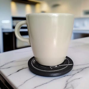 May include: A cream-colored ceramic mug with a curved handle sits on a black coaster with a white design. The coaster is round and has a raised edge. The mug is on a white marble countertop.